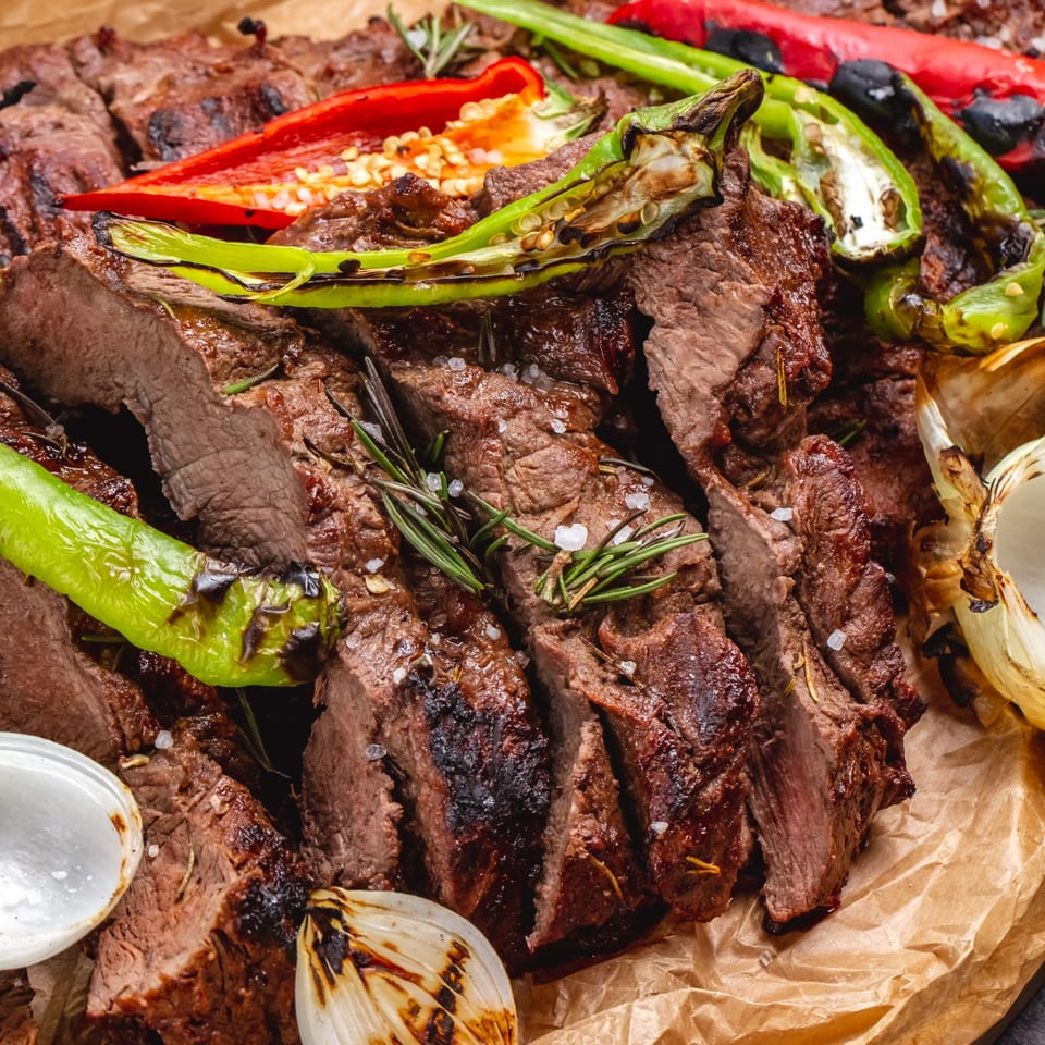 Sliced grilled steak with roasted green and red peppers, onions, and rosemary on parchment paper.