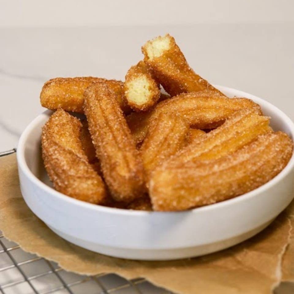 A white bowl filled with golden-brown churros, coated in cinnamon sugar, on a cooling rack.