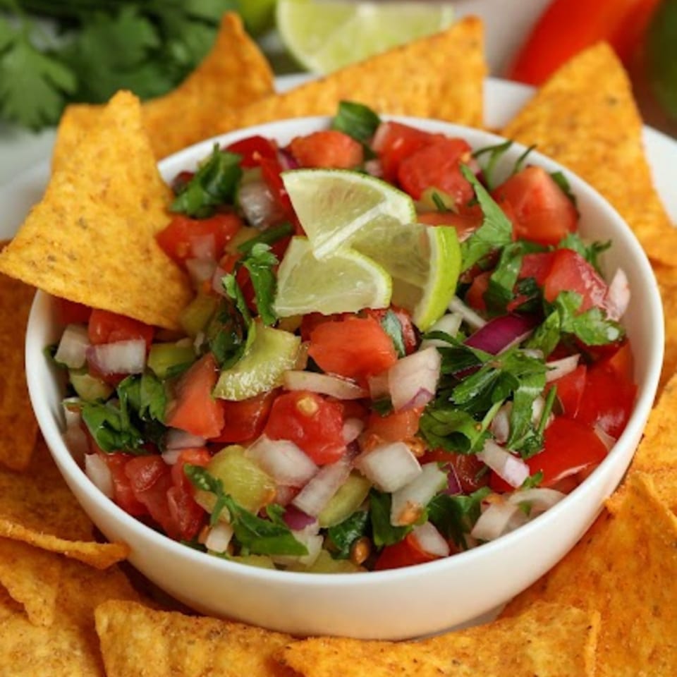 A white bowl of fresh La Estrella Taqueria pico de gallo with lime wedges, surrounded by tortilla chips.