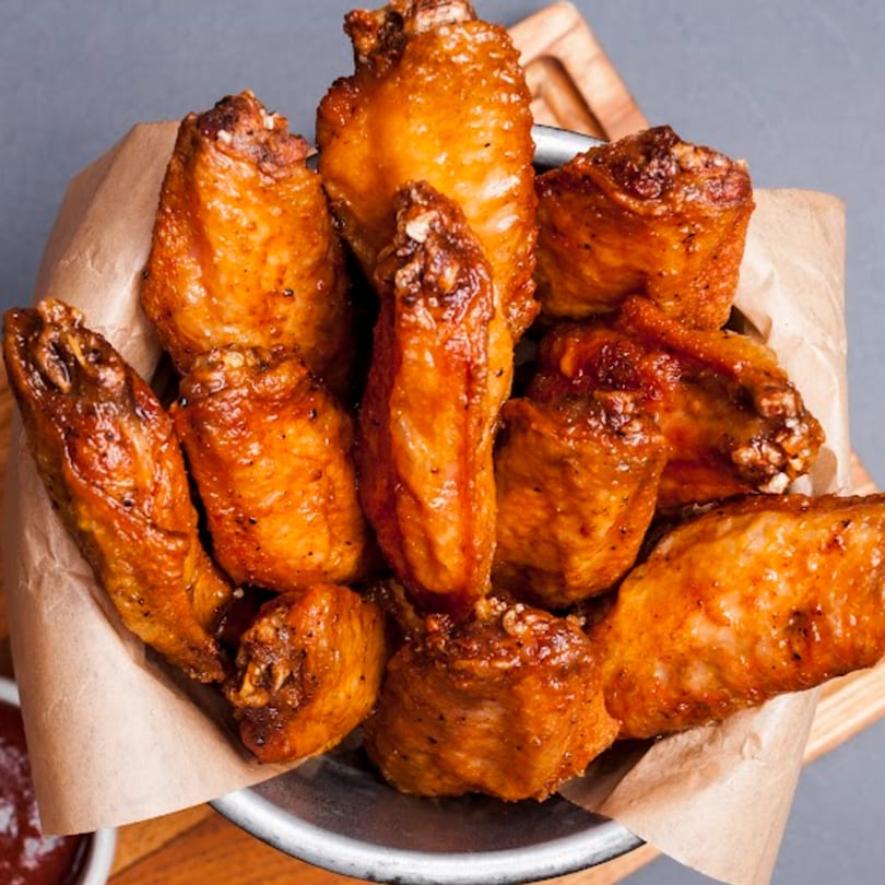 A metal bowl filled with crispy, golden-brown chicken wings, lined with brown parchment paper.